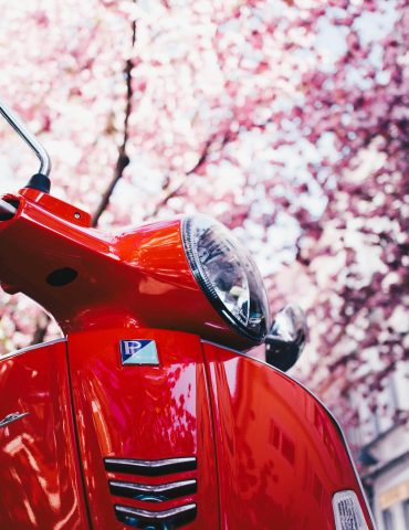 Close-up of a red Vespa scooter parked under blooming trees in a town setting.