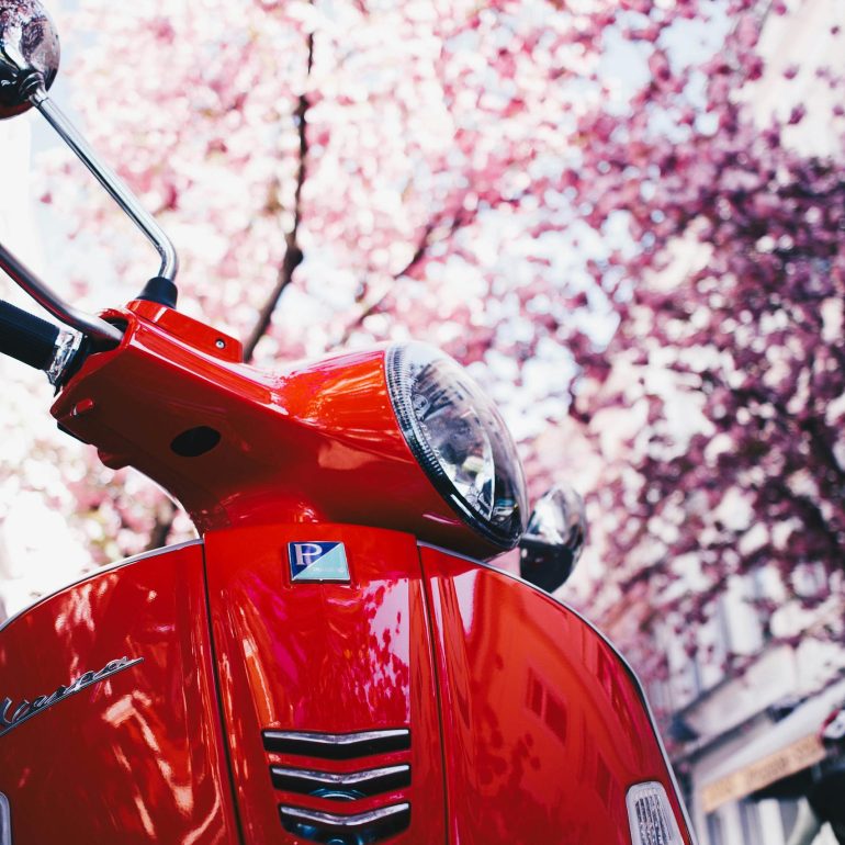 Close-up of a red Vespa scooter parked under blooming trees in a town setting.